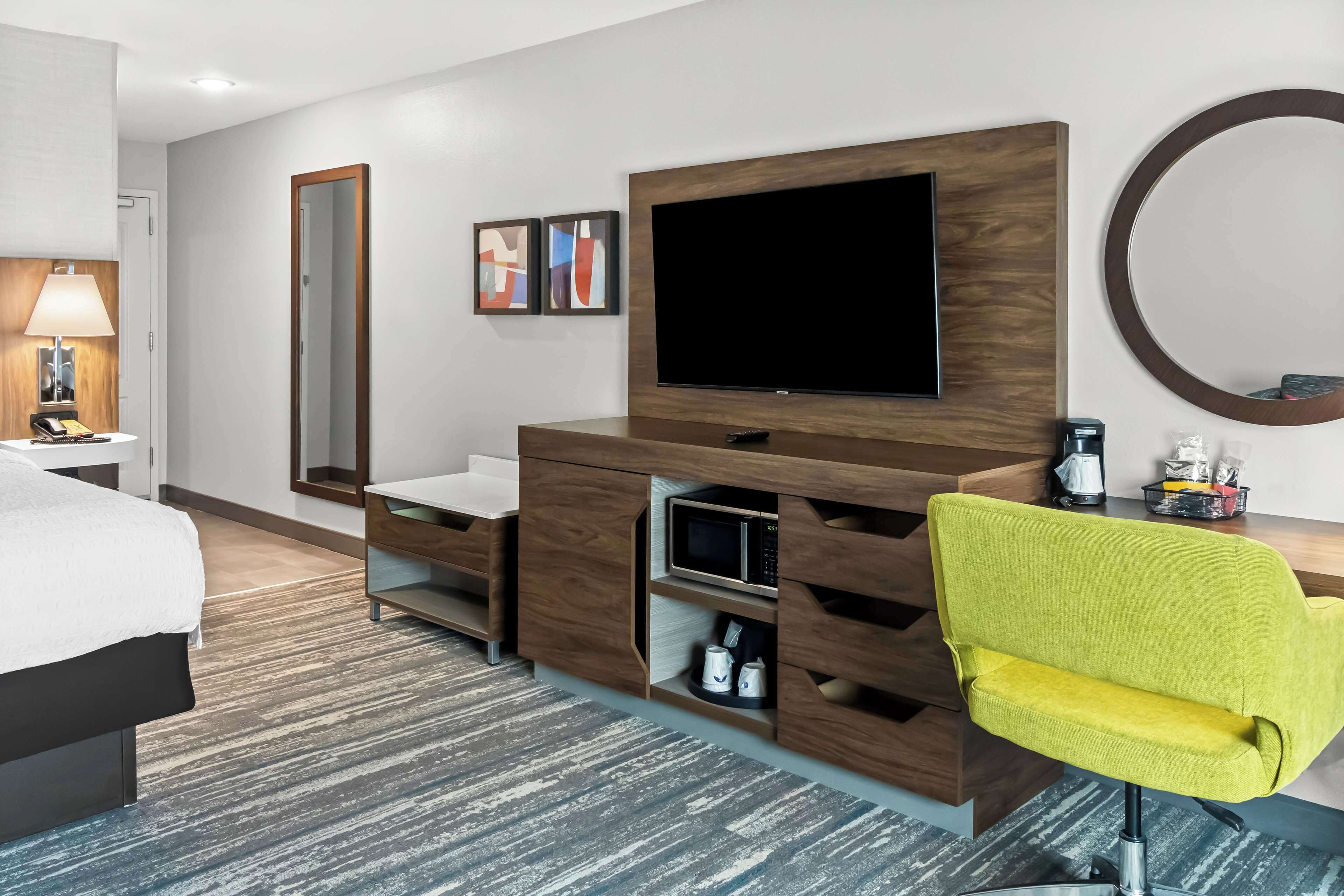 Habitación de hotel con cama, escritorio con televisor de pantalla plana, espejo y una silla amarilla brillante.