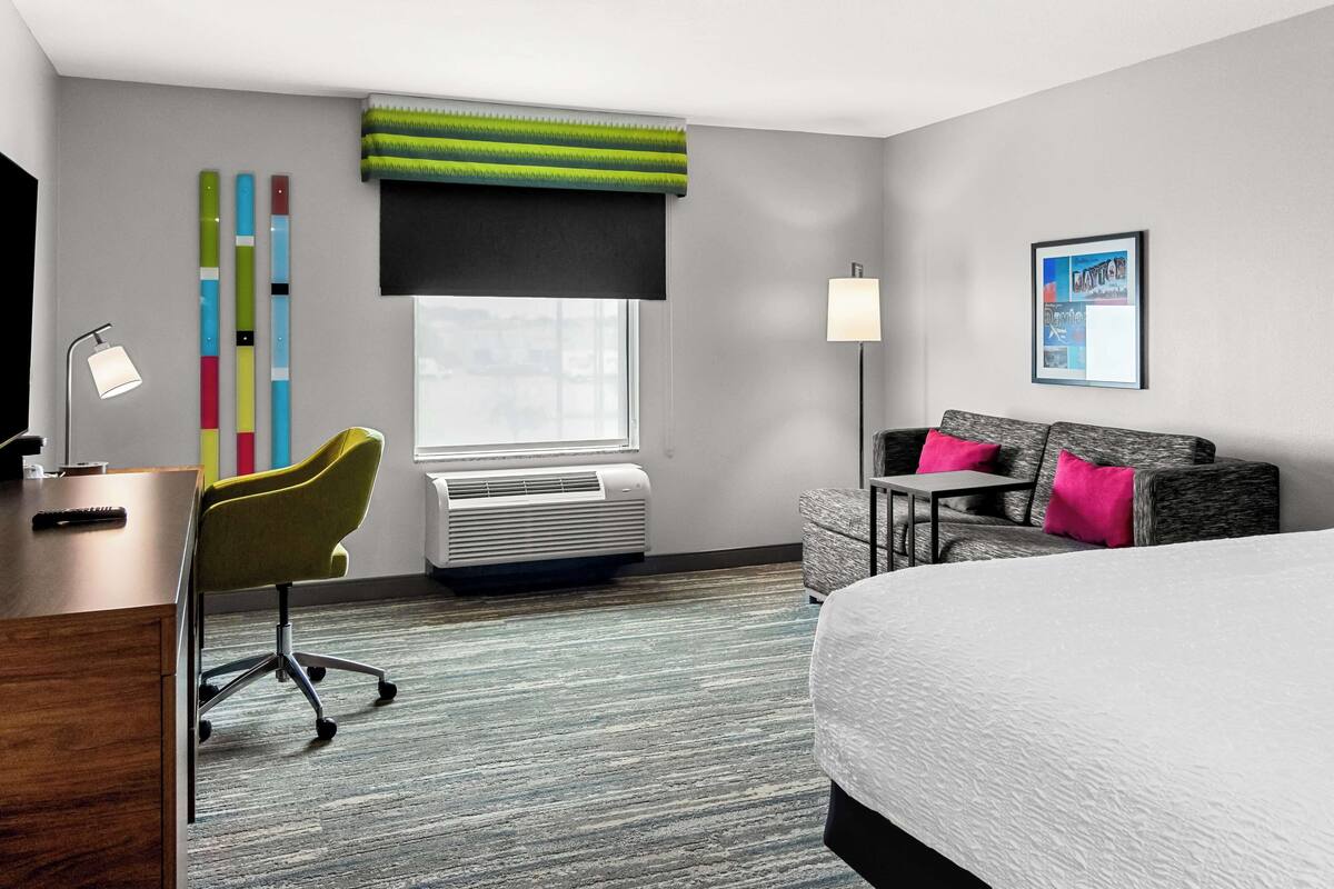 A hotel room with a bed, a desk with a lamp, a chair, a sofa, a window with blinds, a wall-mounted air conditioner, and a framed picture.