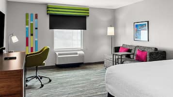 Habitación de hotel con cama, escritorio con lámpara, una silla, sofá, ventana con persianas, aire acondicionado de pared y un cuadro enmarcado.