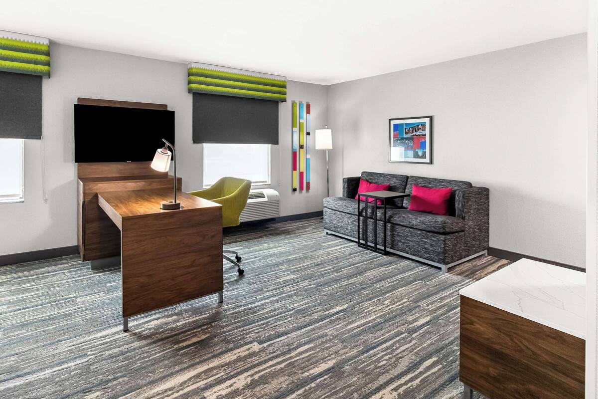 A modern hotel room with a wooden desk, a yellow chair, a grey sofa with pink pillows, a flat-screen TV, and a colorful abstract wall art piece.