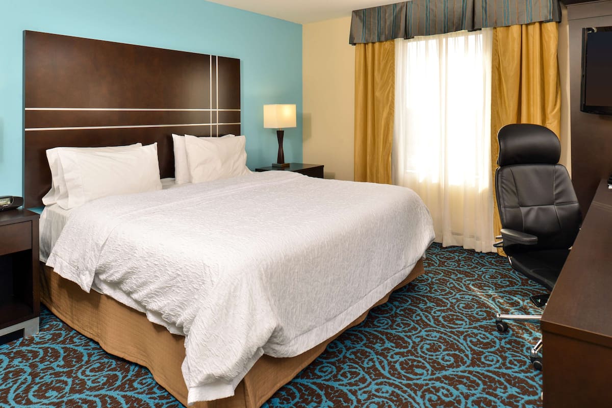 A hotel room with a large bed, a desk, a chair, a nightstand, a lamp, and a window with curtains.