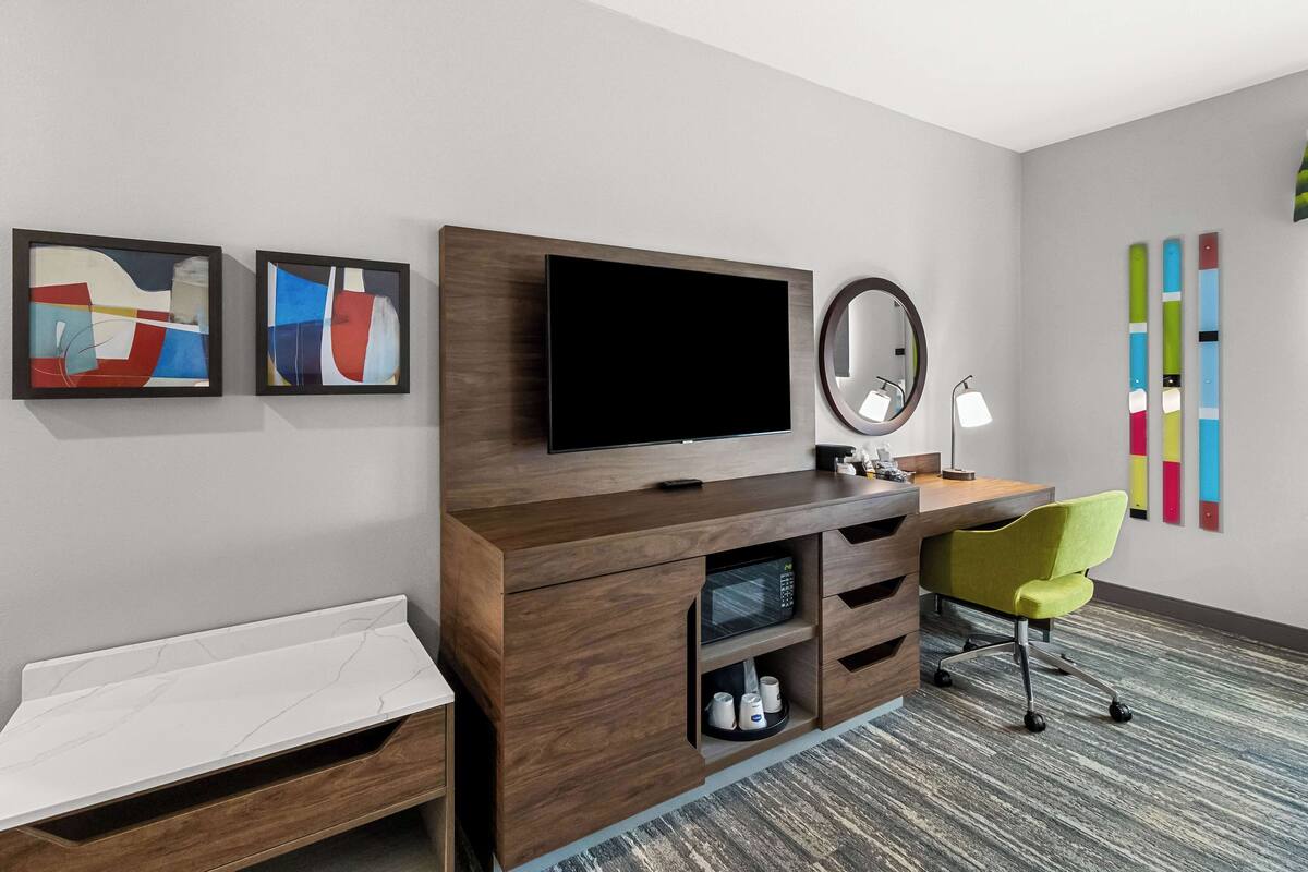 A modern hotel room with a wooden vanity, a flat-screen TV, a mirror, a desk with a lamp, a green chair, and abstract art on the wall.