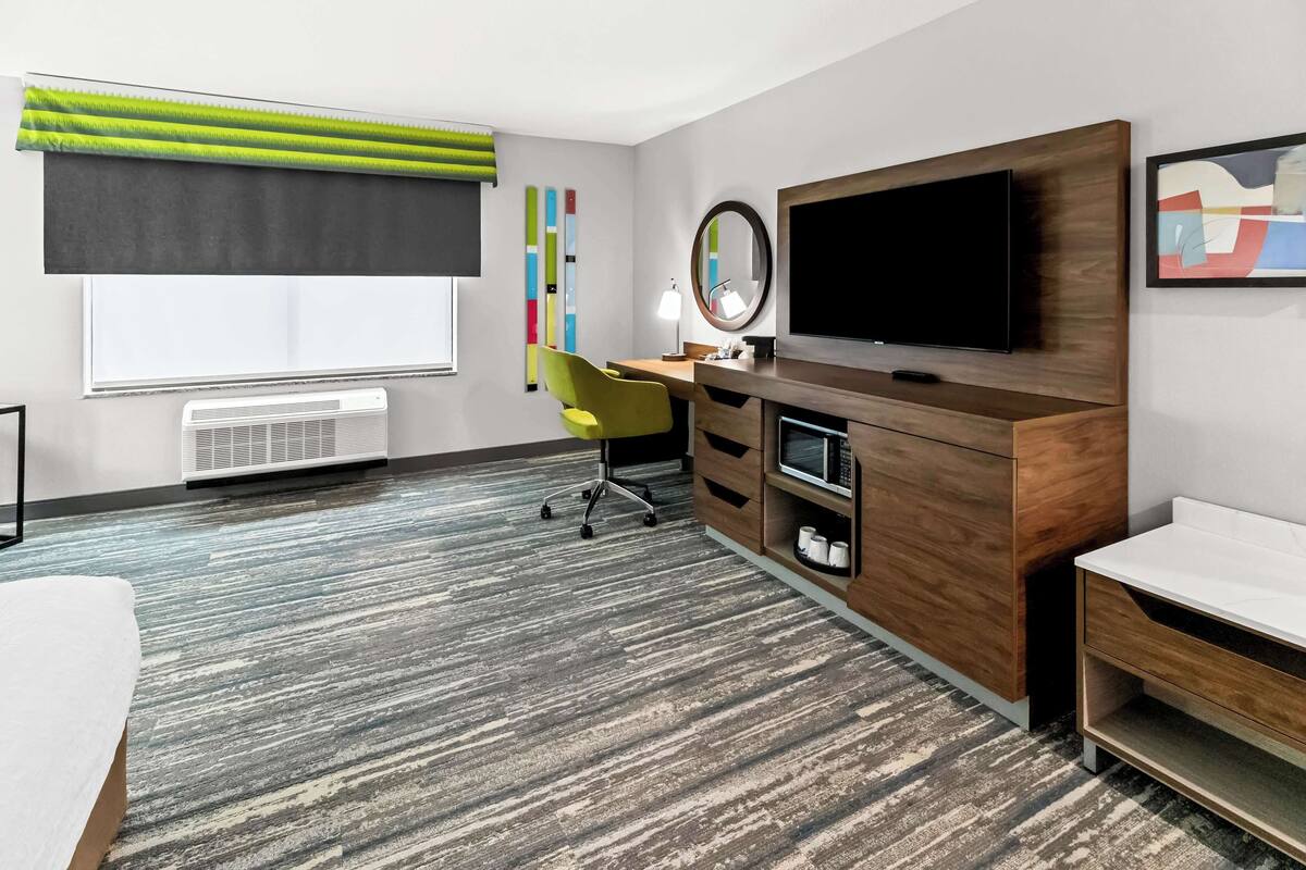 A hotel room with a large flat-screen TV, a wooden desk, a yellow chair, a white bedside table, and a large window with a green and black curtain.