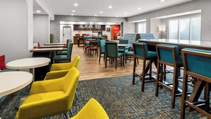A hotel dining area with multiple tables and chairs, a kitchen in the background, and a view of the city through large windows. - Hampton Inn by Hilton Dayton South (Dayton)