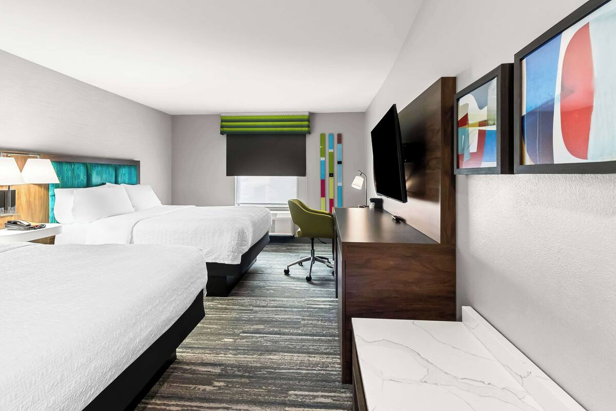 A hotel room with two beds, a desk with a chair, a TV, and artwork on the walls.