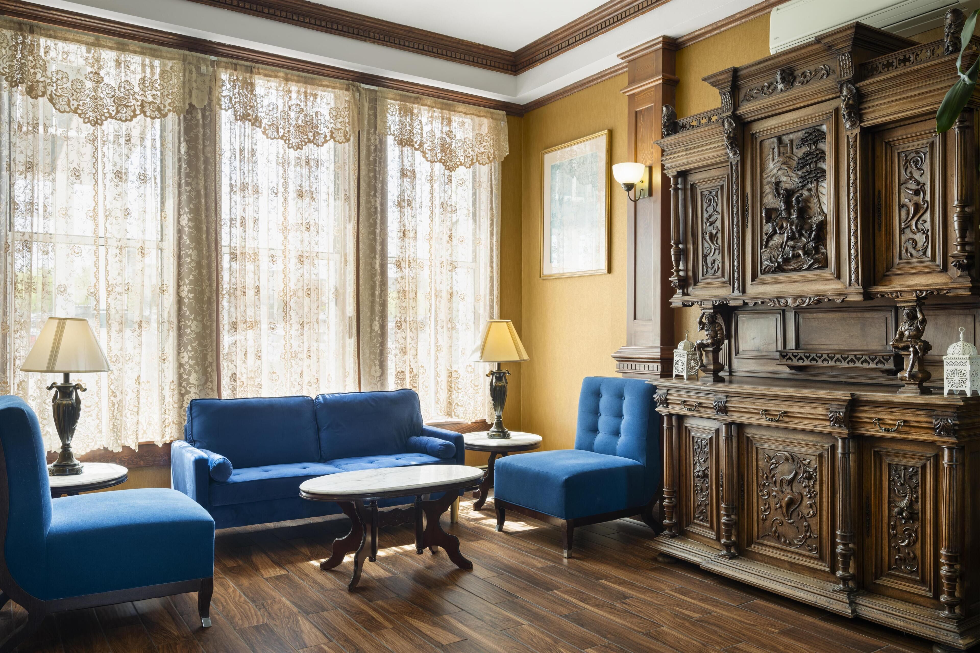 Una habitación con un sofá azul, dos sillones, un aparador de madera con adornos y un ventanal con cortinas de encaje.