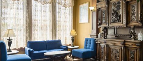 Une pièce avec un canapé bleu, deux fauteuils, un buffet en bois orné de sculptures et une grande fenêtre avec des rideaux en dentelle.