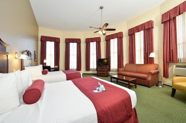 Habitación de hotel con dos camas, un sofá, televisor, ventilador de techo y ventanas grandes con cortinas.
