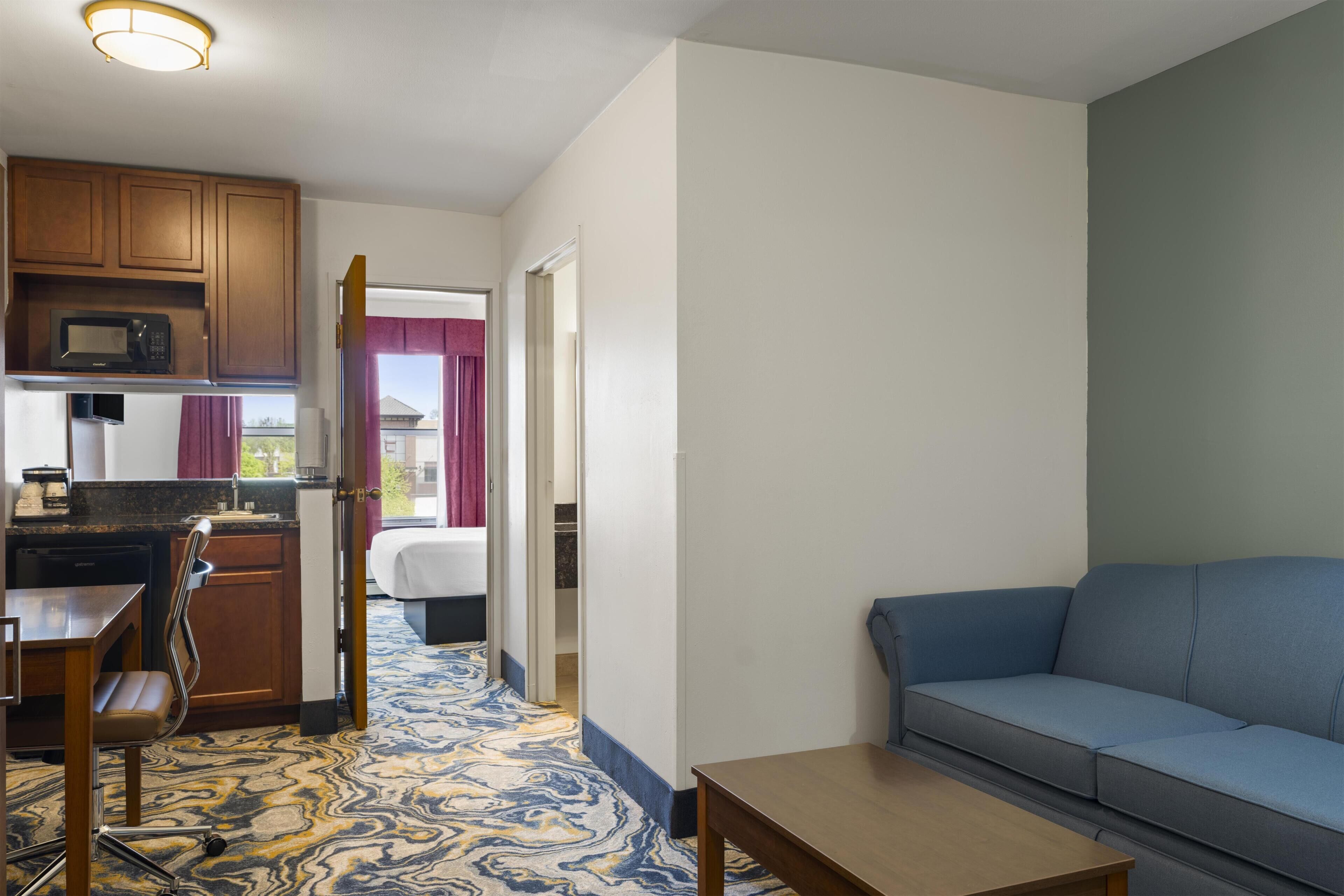 A compact hotel room with a kitchenette, a bed, a sofa, and a patterned carpet.