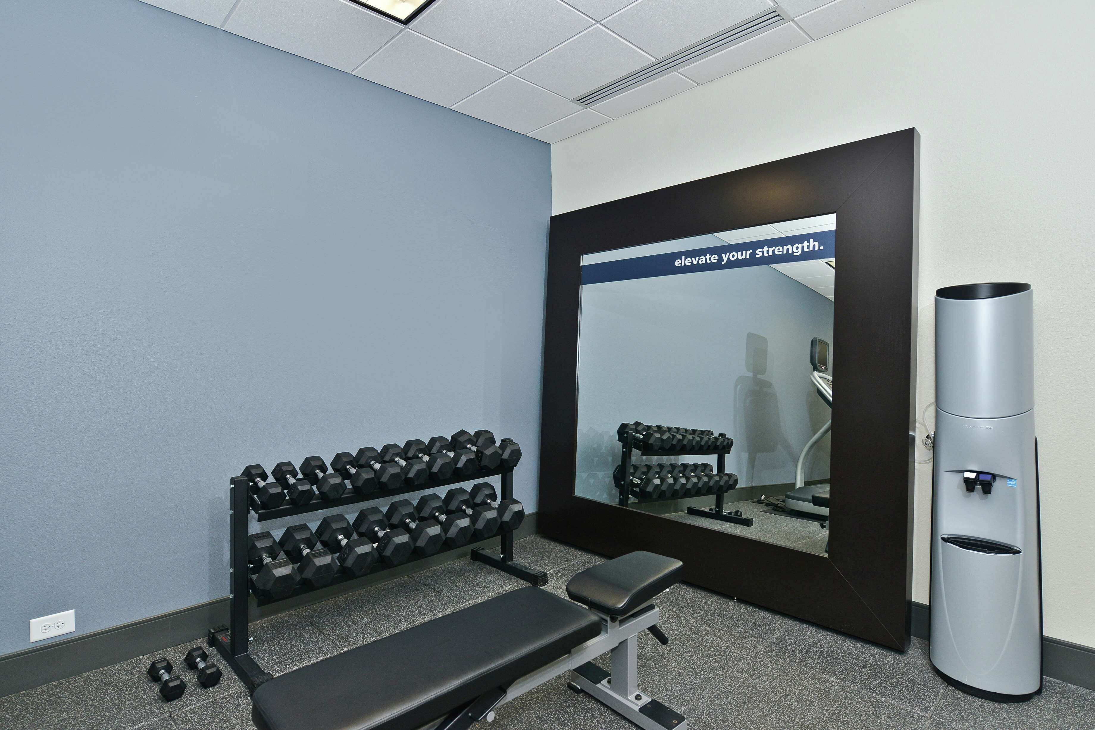 A gym with a bench, dumbbells, a water dispenser, and a motivational mirror.