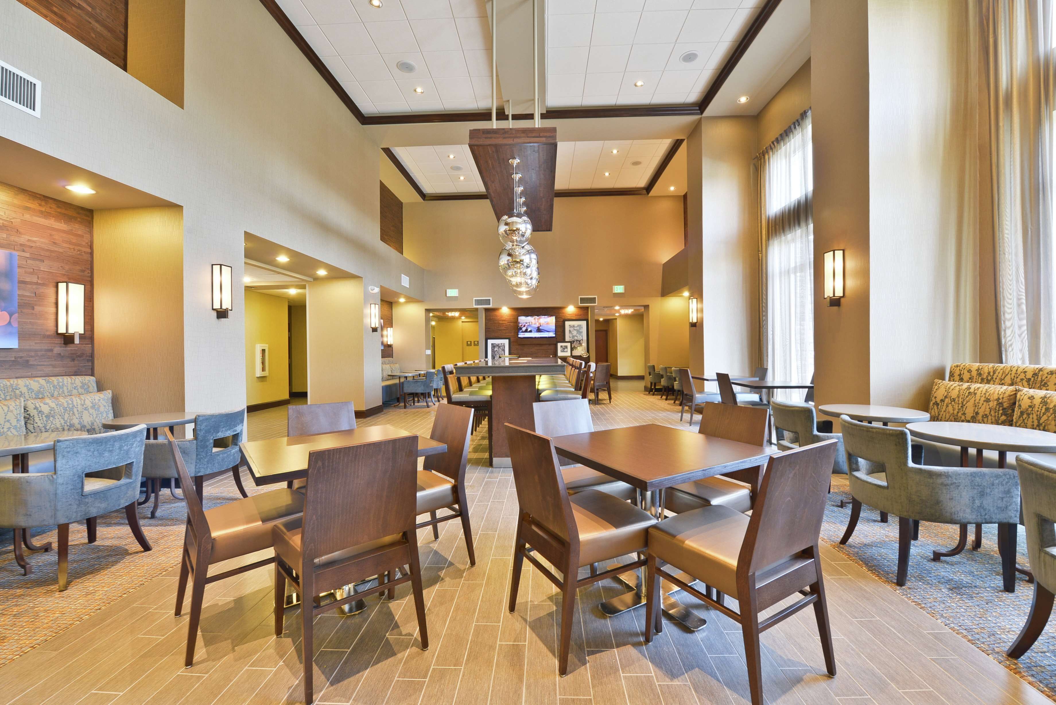 A hotel lobby with multiple seating areas, including tables and chairs, a central light fixture, and a television screen.