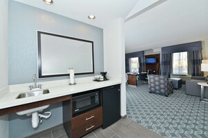 Habitación de hotel con kitchenette, un espejo grande, televisor de pantalla plana y zona de estar con alfombra estampada.