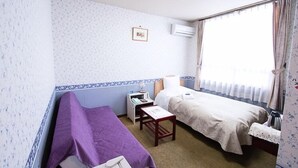 Une petite chambre avec un lit simple, une couverture de lit violette, une table de chevet avec une lampe et une fenêtre avec des rideaux.