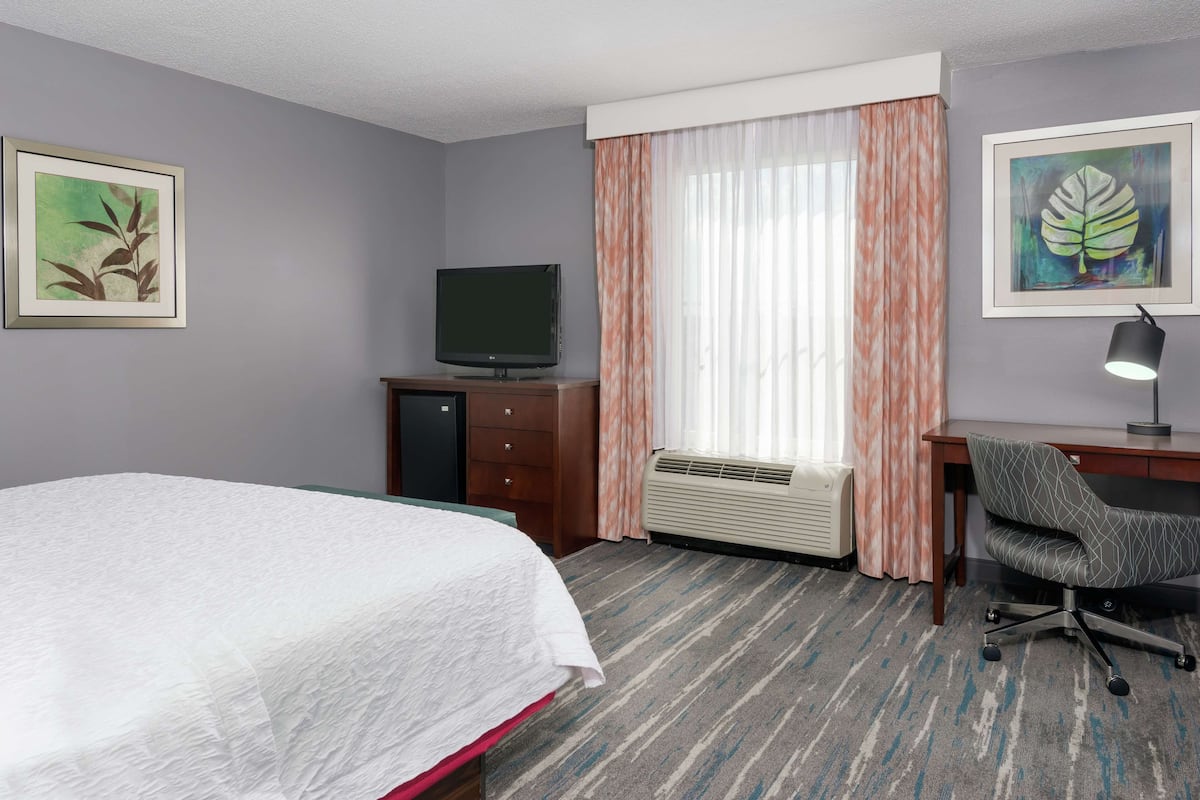 A hotel room with a bed, a desk with a chair, a television, and two framed artworks on the walls.