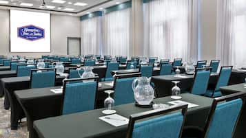 Un salón de conferencias con filas de mesas y sillas, una pantalla grande que muestra el logotipo de Hampton Inn & Suites y un proyector.