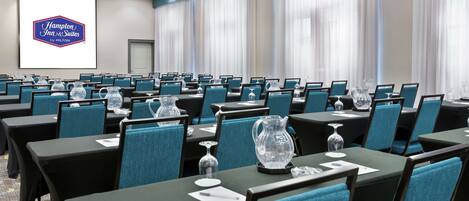 Un salón de conferencias con filas de mesas y sillas, una pantalla grande que muestra el logotipo de Hampton Inn & Suites y un proyector.