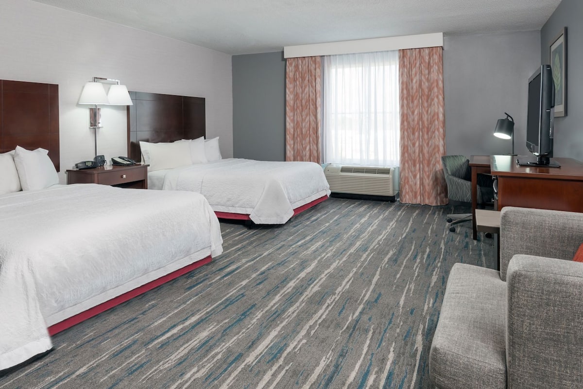 A hotel room with two beds, a desk, a chair, a TV, a lamp, and a window with curtains.
