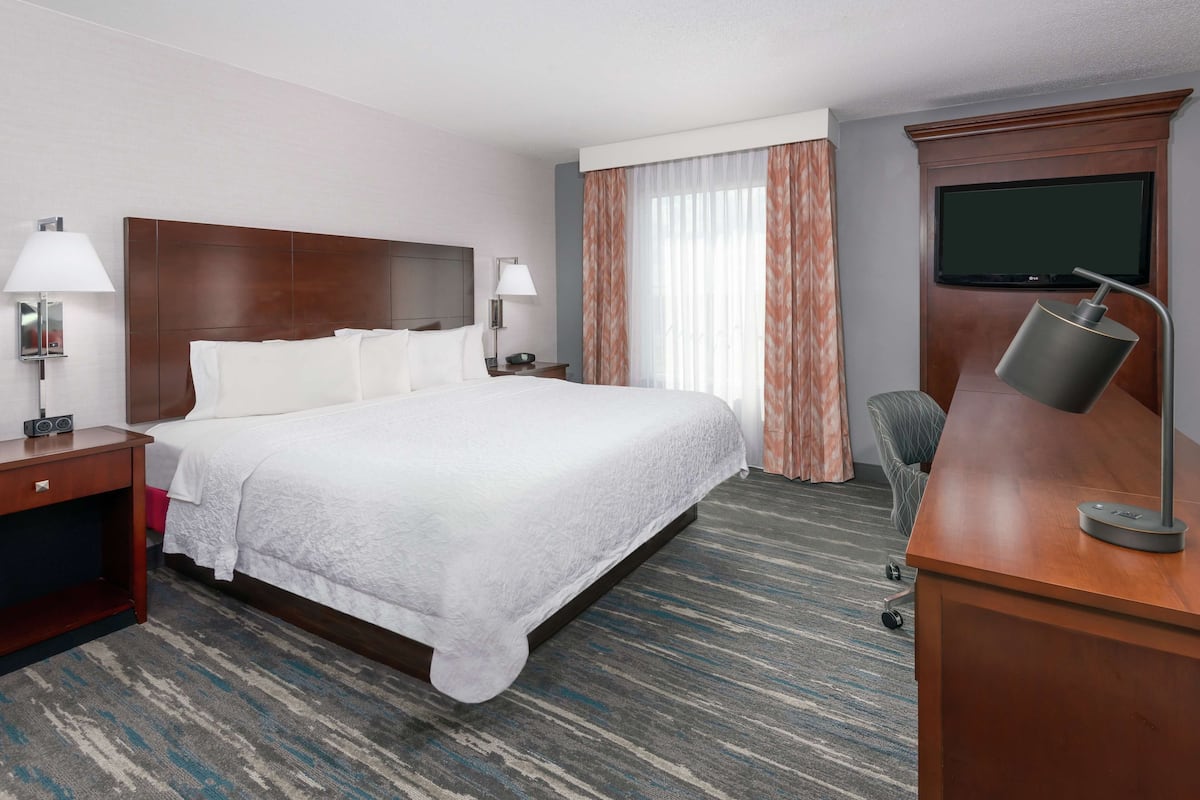A hotel room with a large bed, two bedside lamps, a desk with a computer, a TV mounted on the wall, and a window with curtains.