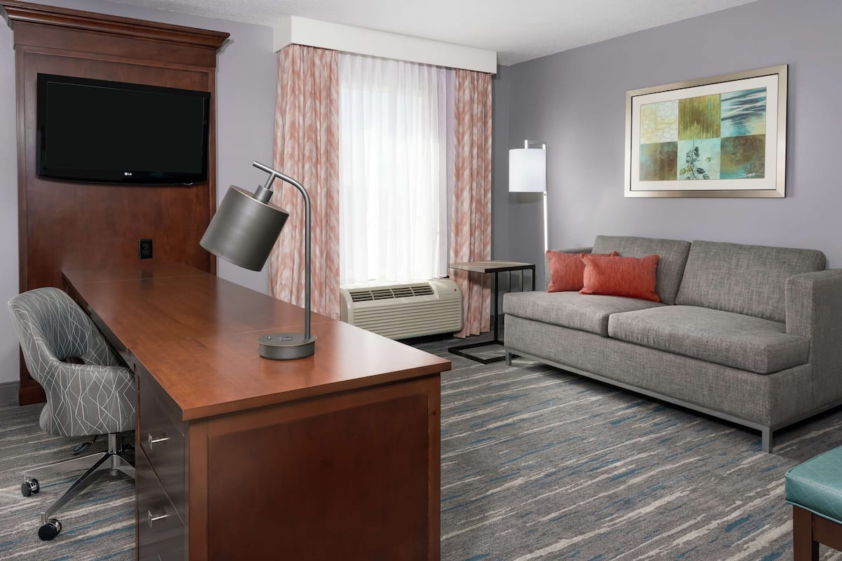 A hotel room with a wooden desk, a grey sofa, a TV mounted on the wall, and a framed artwork.