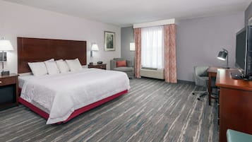 Habitación de hotel con una cama grande, un escritorio con televisor, una silla, una lámpara de escritorio, una ventana con cortinas y un equipo de aire acondicionado.