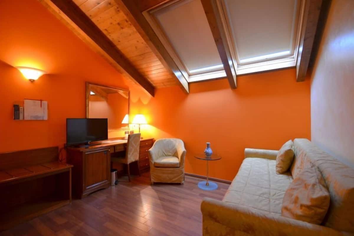 A room with a wooden ceiling, a television, a desk with a lamp, a chair, a sofa, and a small table with a vase.