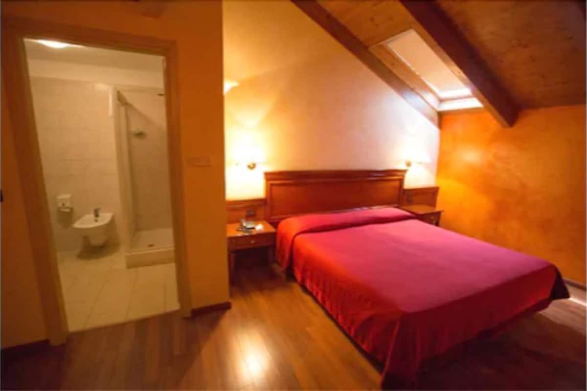 A bedroom with a wooden floor, a bed with a red bedspread, a wooden headboard, and a nightstand with a lamp.