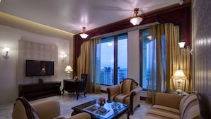 Habitación de hotel con televisor de pantalla plana, sofá, sillones, una mesa de centro y un amplio ventanal con cortinas.