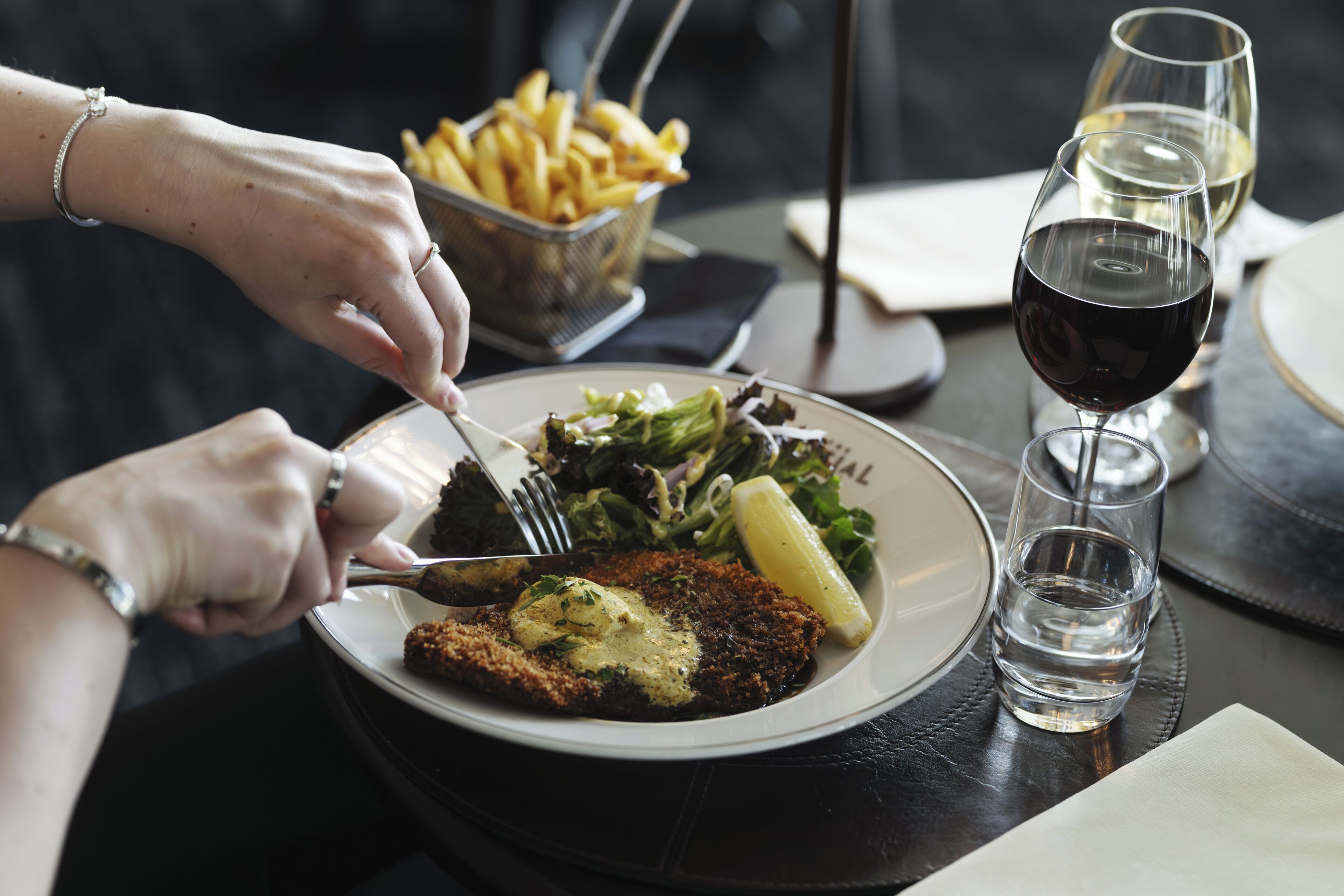 Uma mesa de jantar com um prato de comida, um copo de vinho tinto e um cesto de batatas fritas.