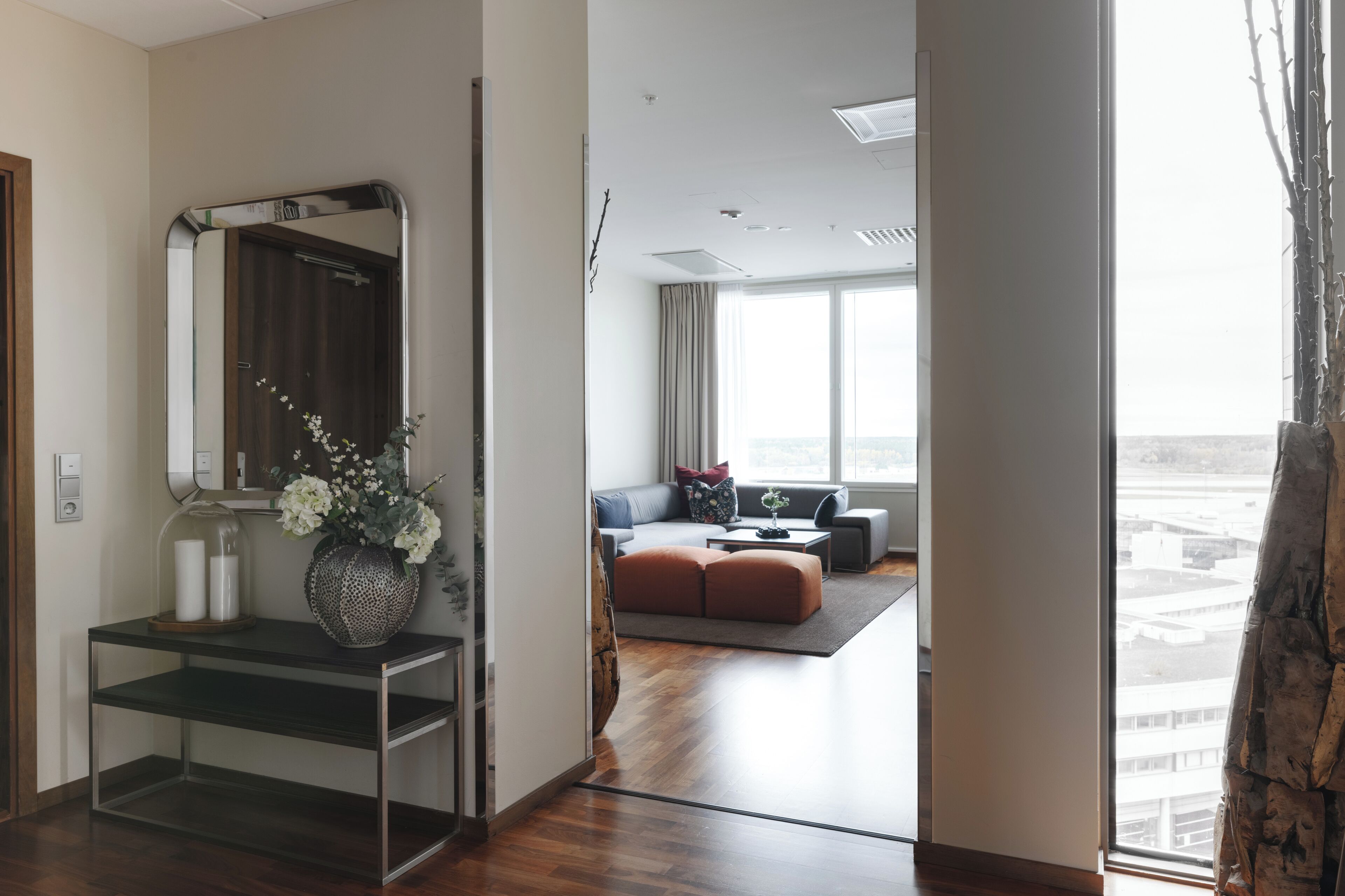 A hotel room with a wooden floor, a sitting area with a sofa and ottoman, a large window with curtains, and a mirror with a vase of flowers.