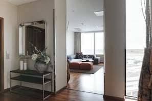 A hotel room with a wooden floor, a sitting area with a sofa and ottoman, a large window with curtains, and a mirror with a vase of flowers.