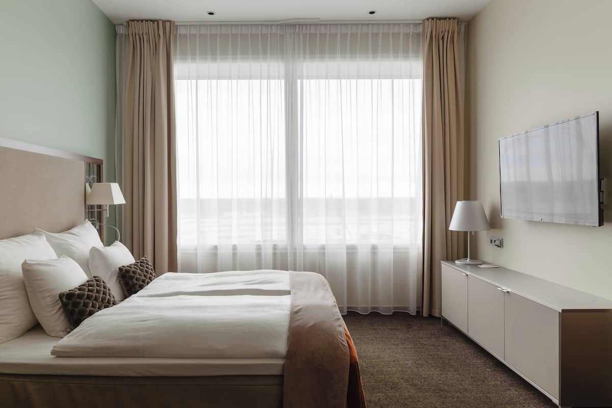 A hotel room with a large bed, two bedside tables with lamps, a TV on the wall, and a view through sheer curtains.