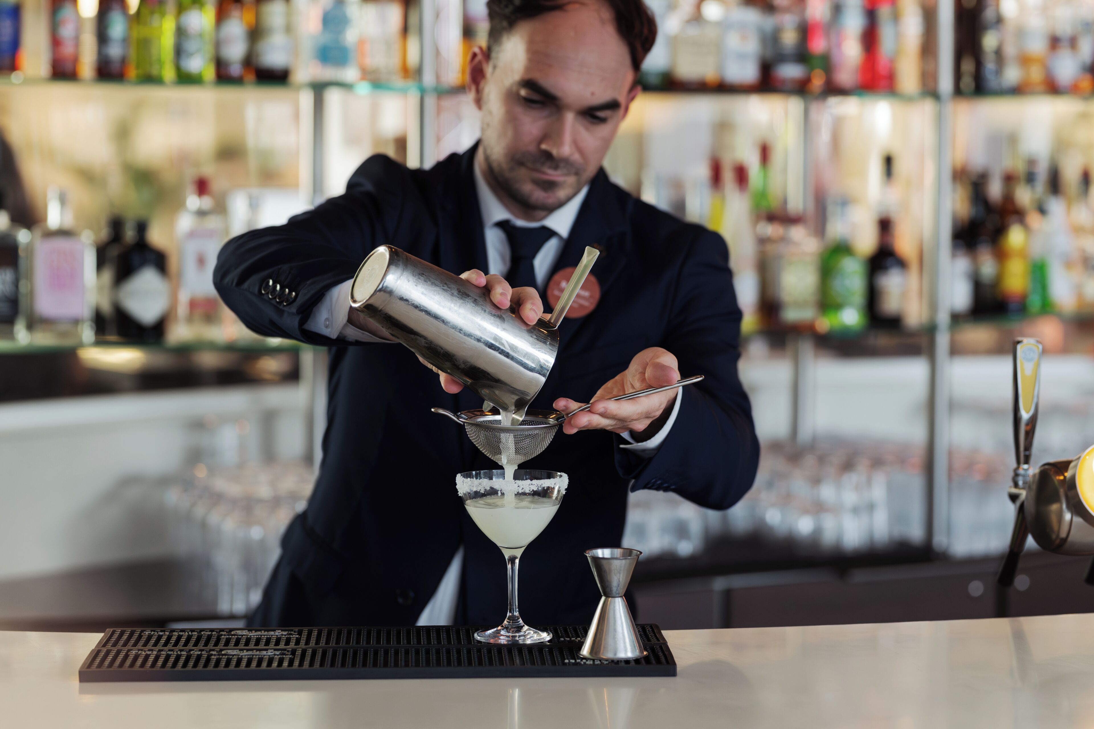 Un cantinero preparando una bebida en la barra, con estantes llenos de botellas al fondo.