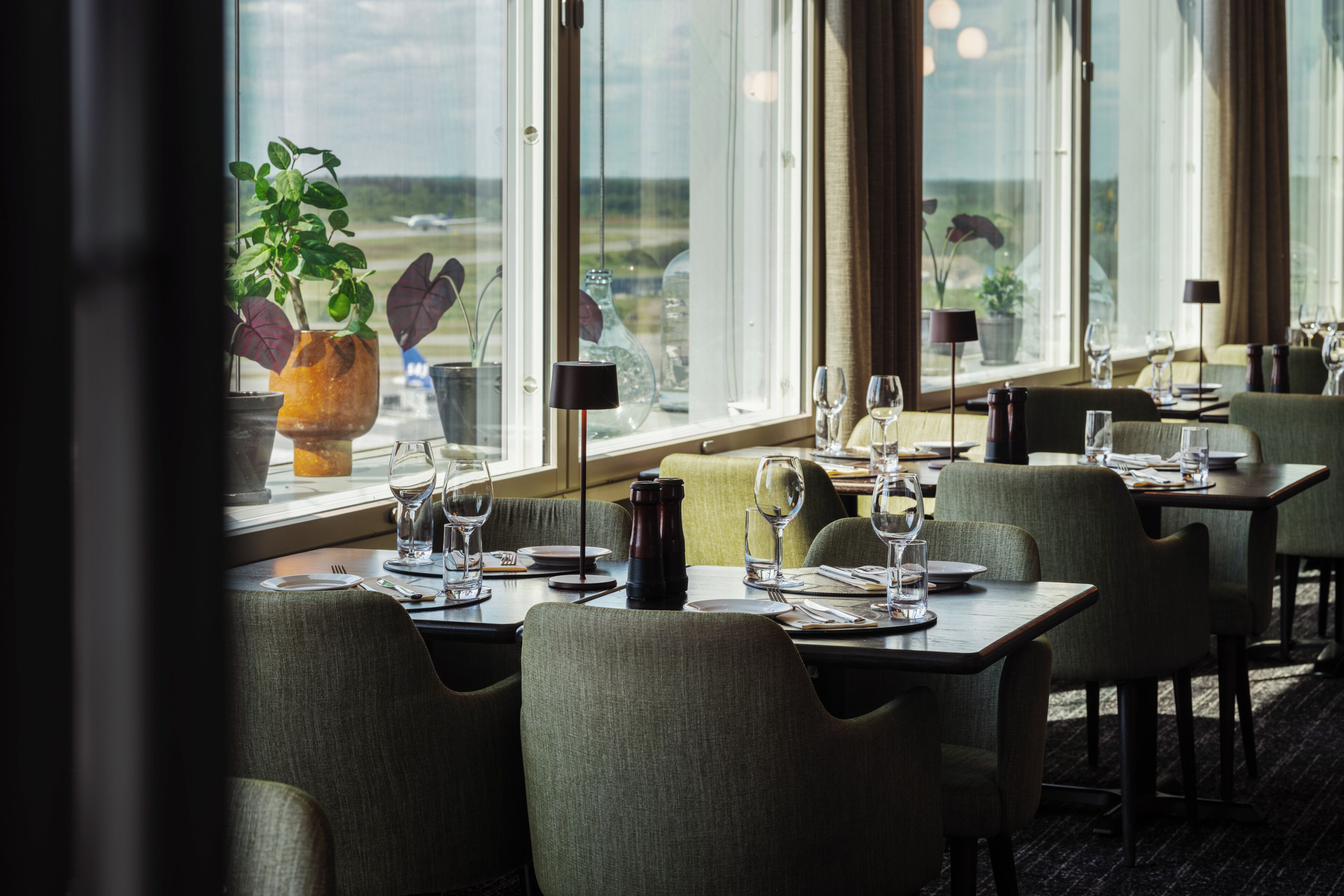 A restaurant with tables set for dining, large windows offering a view of the airport, and potted plants on the windowsill.