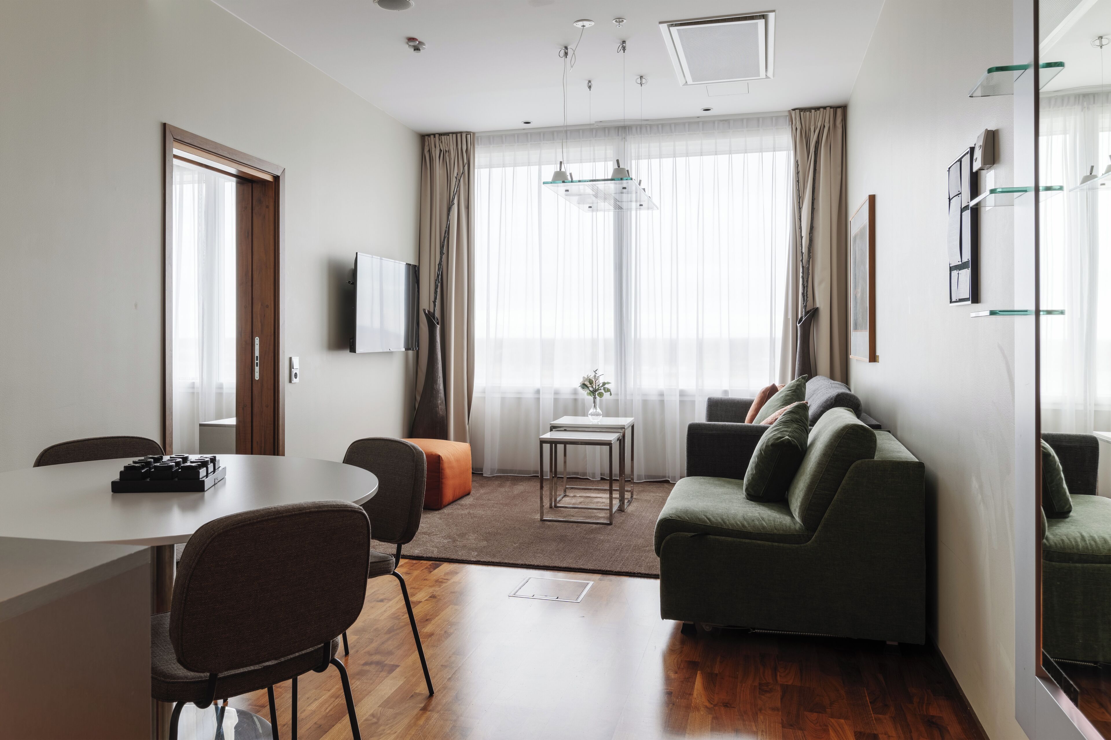 A modern living room with a sofa, a dining table with chairs, a TV mounted on the wall, and a large window with curtains.