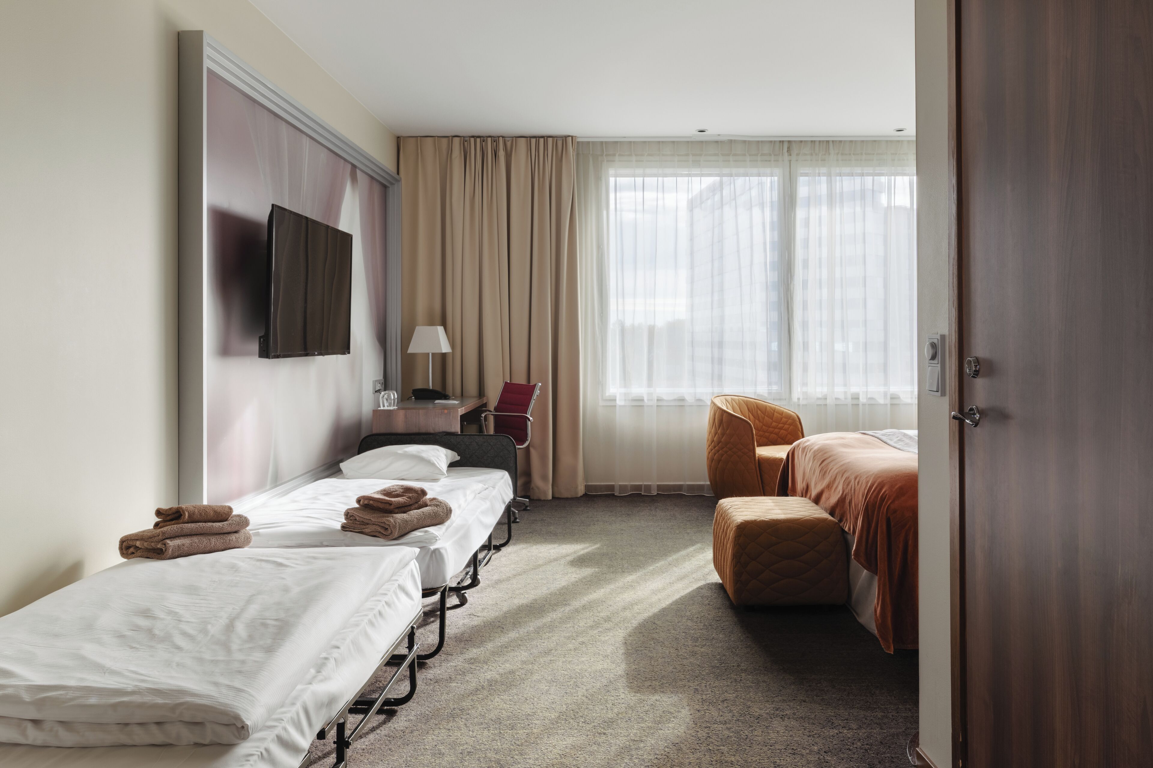 Habitación de hotel con dos camas, televisión, escritorio con lámpara, una silla y un ventanal grande con cortinas.