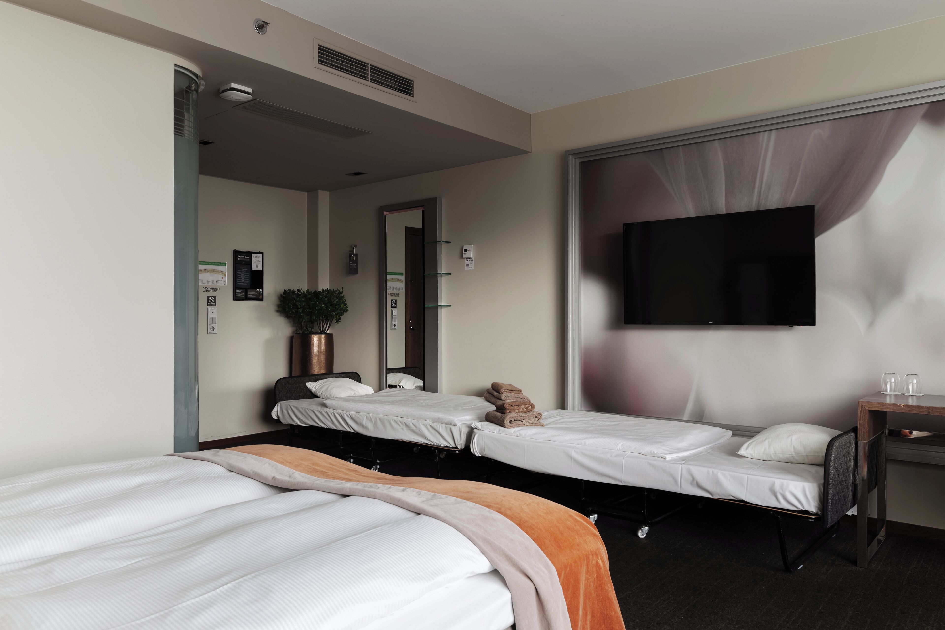 A hotel room with two single beds, a desk with a chair, a flat-screen TV mounted on the wall, and a potted plant.