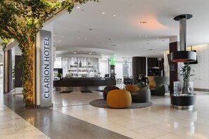 A modern hotel lobby with a reception desk, seating areas, and a decorative plant arrangement.