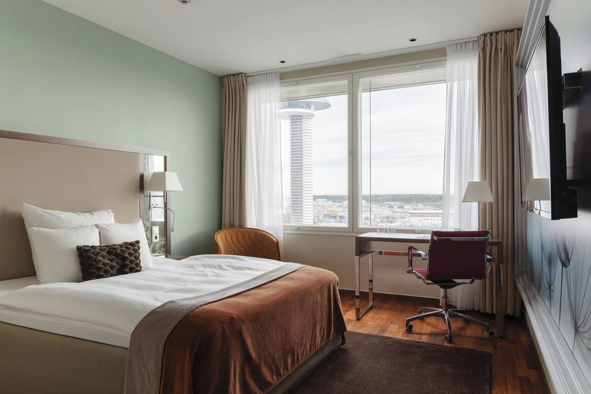 A hotel room with a bed, a desk, a chair, and a large window with a city view.