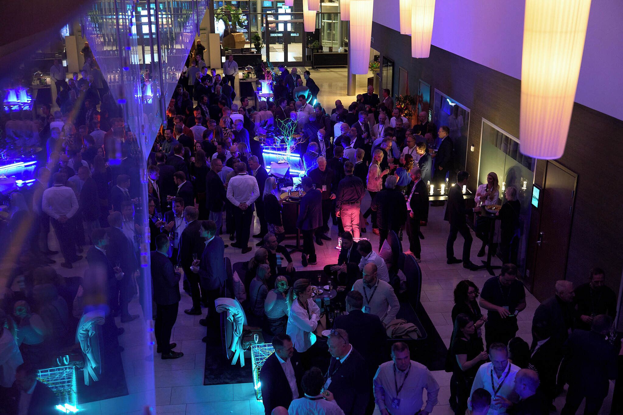 A crowded indoor event with people mingling, tables with drinks, and a modern interior with hanging lights.