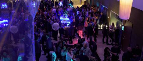 A crowded indoor event with people mingling, tables with drinks, and a modern interior with hanging lights.