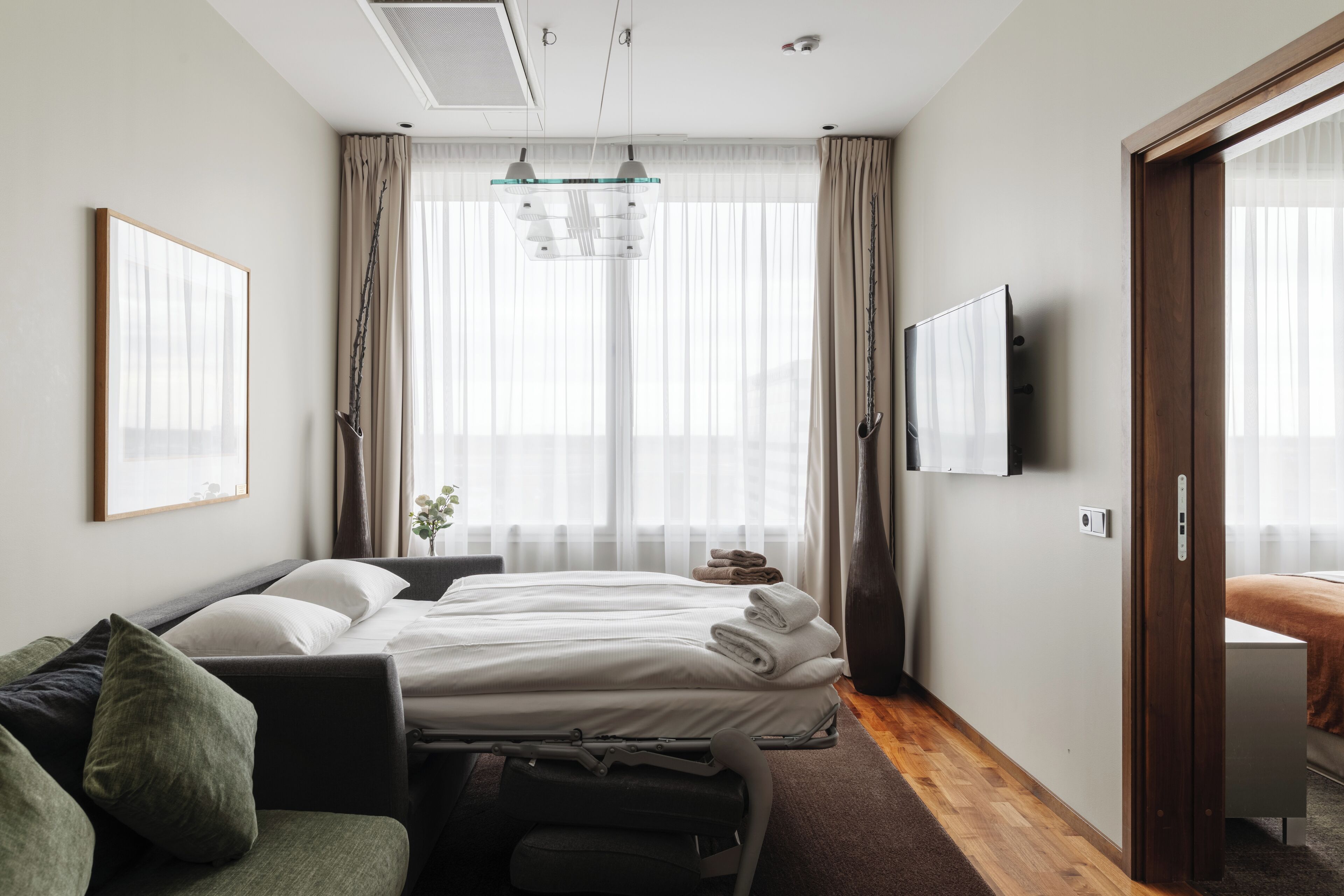 A modern hotel room with a bed, a sofa, a TV, and a large window with curtains.