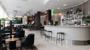 Zona de bar moderna con una barra curva, taburetes y una variedad de botellas. Hay áreas de estar con mesas y sillas, y el espacio está bien iluminado con luces de techo.