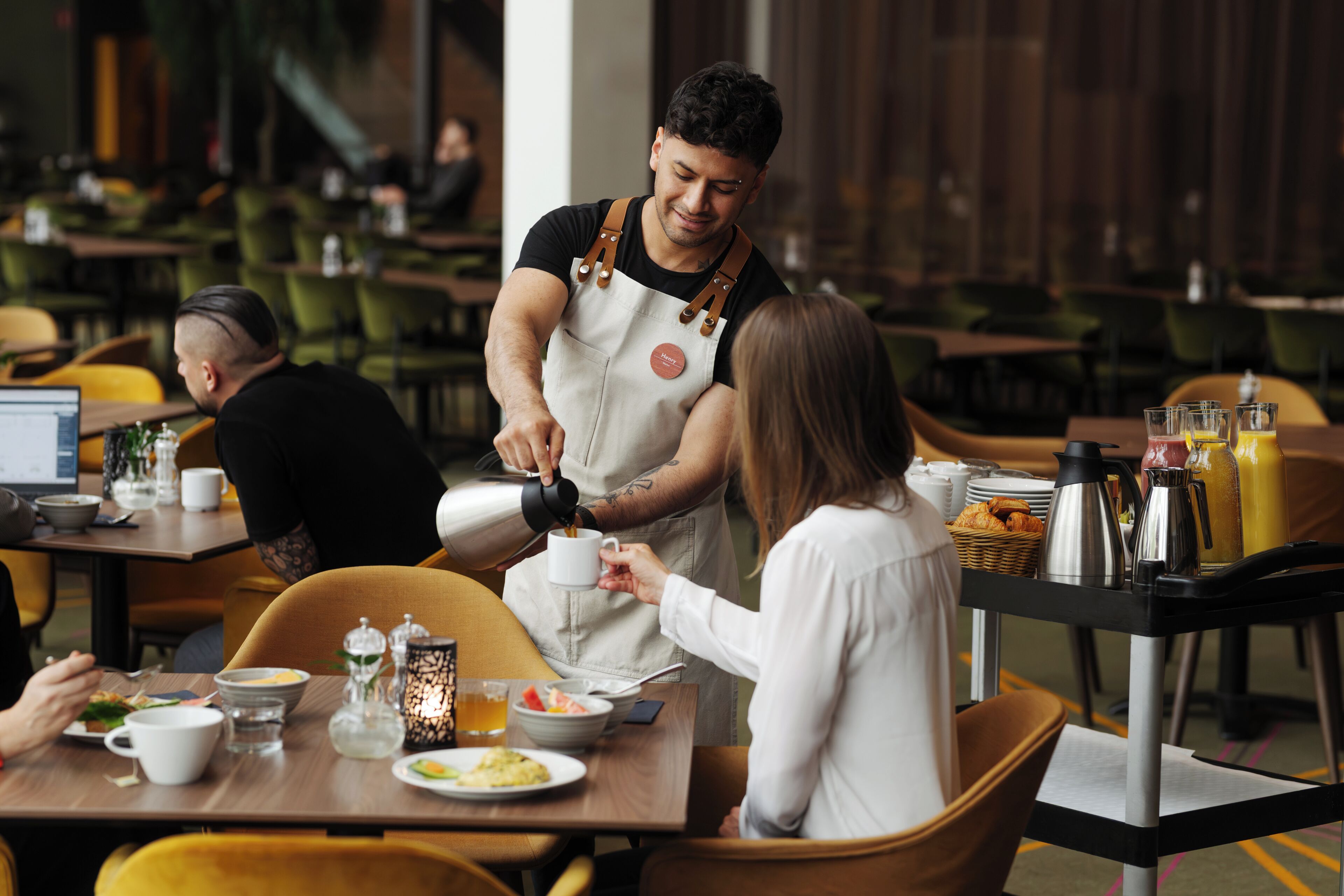 A restaurant setting with a waiter serving coffee to a customer, dining tables with chairs, and various food and drink items on the tables.