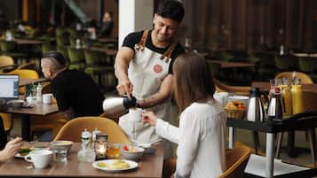 A restaurant setting with a waiter serving coffee to a customer, dining tables with chairs, and various food and drink items on the tables.
