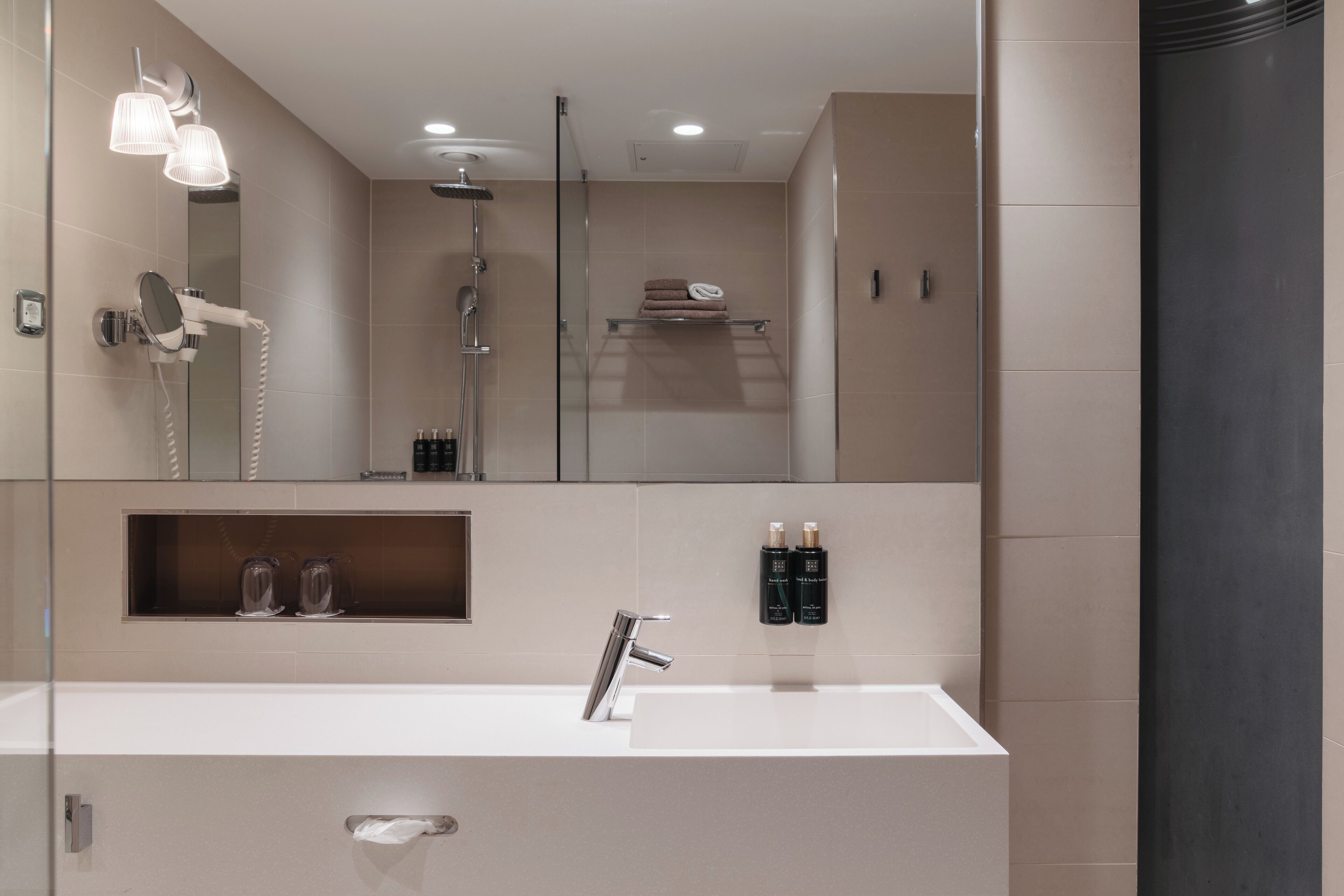 A modern bathroom with a large mirror, a sink, and a shower. There are two bottles of shampoo on the wall.