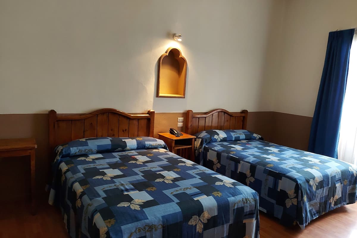 A hotel room with two beds, a bedside table with a phone, a wall-mounted light, and a window with curtains.