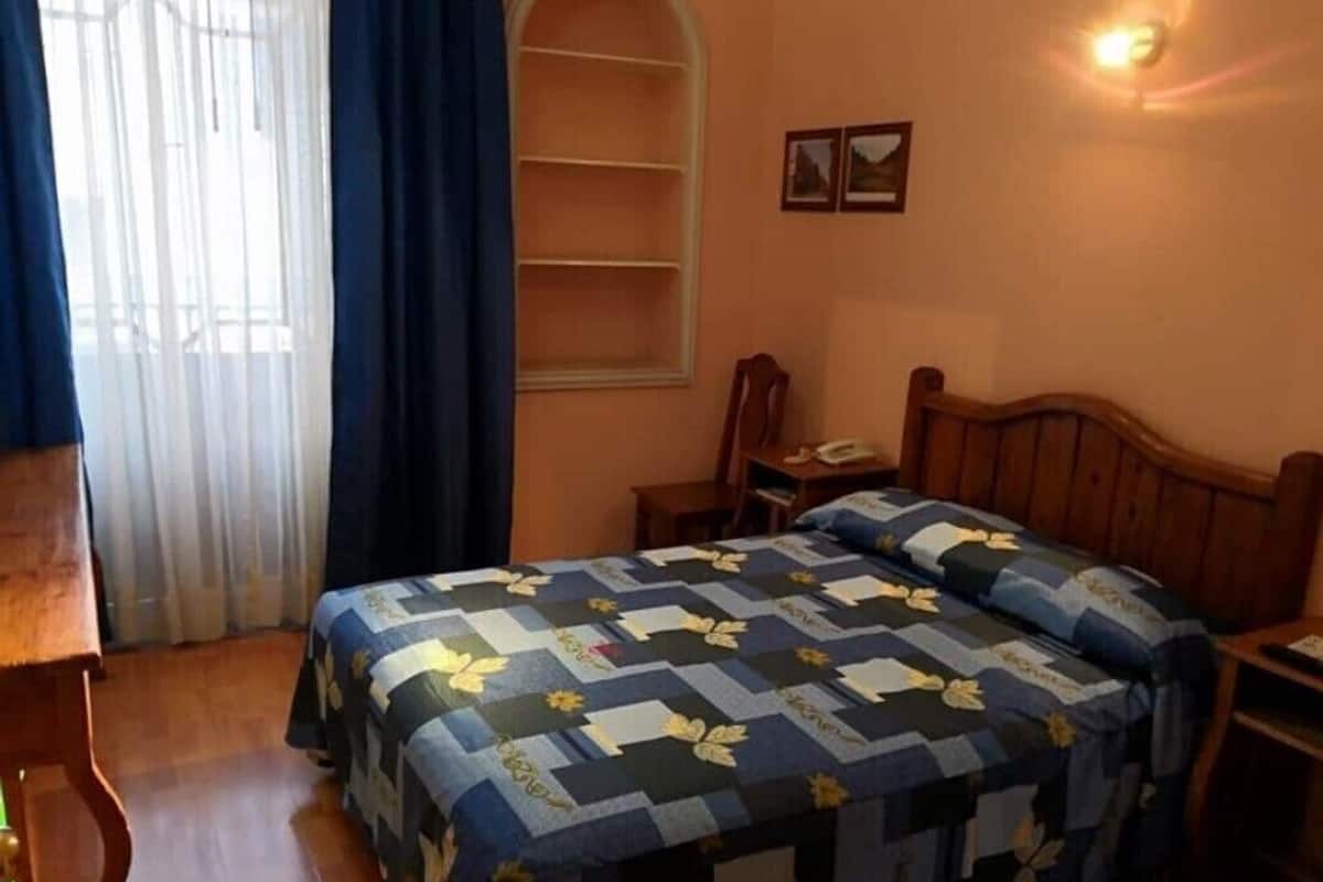 A bedroom with a bed, a nightstand, a chair, a desk, a window with curtains, and a wall-mounted shelf.