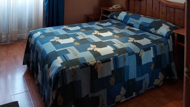 A bedroom with a wooden headboard, a quilted bedspread, a bedside table with a lamp, and a window with curtains. - Hotel de Talavera (Puebla)