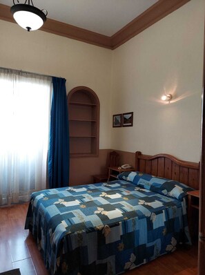 Un dormitorio con cabecera de madera, colcha acolchada, mesita de noche con lámpara y ventana con cortinas.
