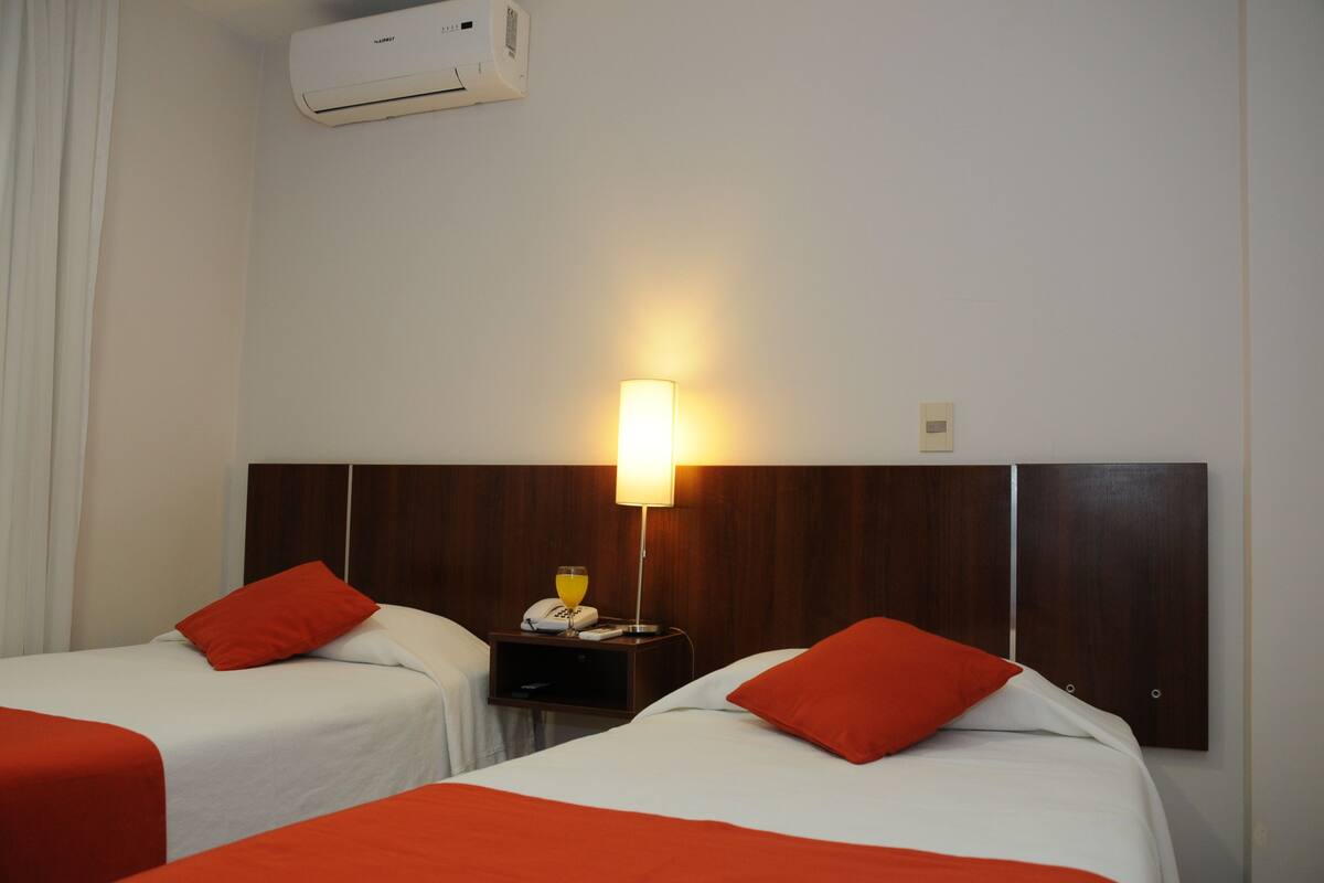 A hotel room with two beds, each with a red pillow and white bedding. There is a nightstand with a lamp, a phone, and a glass of orange juice. An air conditioning unit is mounted on the wall above the beds.